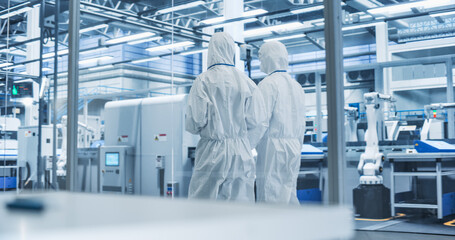 Sterile Modern Factory: Professionals in Coveralls Having a Conversation while Walking Through a Plant. Medical Electronics Manufacturing Laboratory with CNC Machinery and Automated Robotic Arms