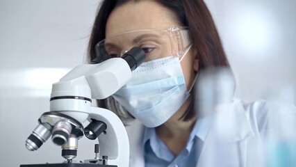 Female scientist in lab goggles and protective mask using microscope in laboratory