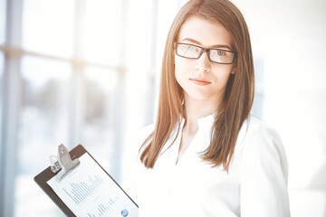 Beautiful female specialist with clipboard standing in sunny office and smiling charmingly. Business people concept