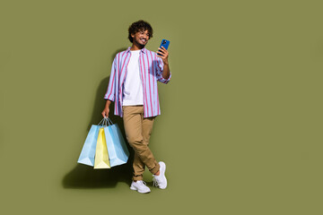 Wall Mural - Full size photo of positive guy dressed striped shirt pants hold shopping bags look at smartphone isolated on khaki color background