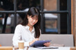 © wattana - Young woman sitting at a table in a cozy, modern café, working intently on a digital tablet with a stylus.