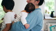 © Marco - Mother holding newborn baby close while another child stands nearby eating. This image captures the busy and loving dynamics of parenthood in a cozy home environment