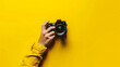 © john - Man's hand holding a camera on a bright yellow background.