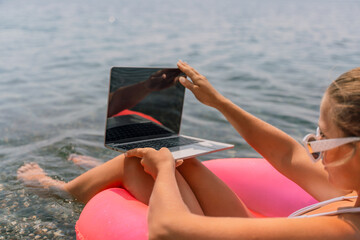 Wall Mural - A woman is sitting in a pink inflatable raft with a laptop on her lap. She is wearing sunglasses and she is enjoying her time by the water.