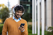 © Liubomir - Young man smiling while looking at smartphone outdoors in urban environment. Wearing orange sweater and headphones, enjoying music. Scene conveys leisure, technology, and happiness in city life.