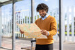 © Liubomir - Happy young man with curly hair exploring city using map, standing at bus stop with suitcase. Captures travel, adventure, and navigation on bright sunny day.