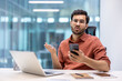© Liubomir - Man in office showing confusion holding smartphone, expressing frustration during work. He is sitting at desk with laptop and tablet, conveying business communication or technology issues.