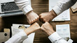 © Nima - Four hands bump in the center of a table, symbolizing collaboration, partnership, teamwork, and agreement. Papers and a laptop are visible, representing the work environment.