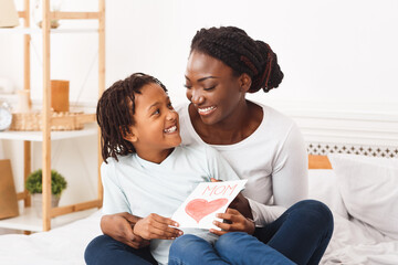 Wall Mural - Congatulations. Happy african mom embracing her little child and holding greeting card, sitting on bed, copyspace