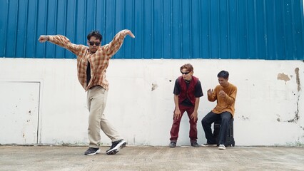 Wall Mural - Young street dancer dancing in hip hop style with multicultural friends cheering behind at wall. Attractive handsome man stretch arms at street with blue background. Outdoor sport 2024. Hiphop.