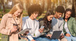 © Prostock-studio - International young friends sitting on bench in park, using gadgets