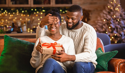 Wall Mural - Romantic Surprise For Christmas. Afro husband covering his wife's eyes with hand and presenting gift while sitting on sofa near Xmas tree