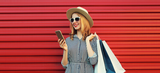 Wall Mural - Beautiful happy young woman looking at phone with shopping bag in summer hat on red background