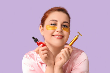 Sticker - Young woman with under-eye patches holding facial massage tool and bottle of cosmetic product on lilac background