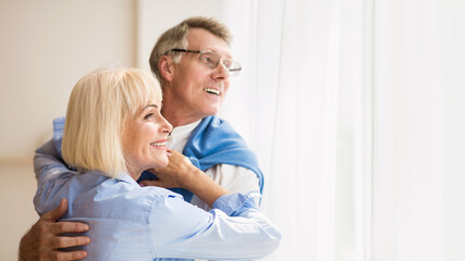 Wall Mural - Happy senior couple looking through window and hugging at home, free space