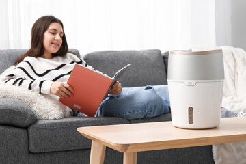 Wall Mural - Young woman reading magazine on sofa near modern humidifier in living room