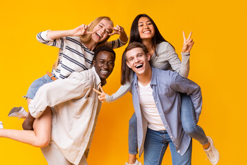 Wall Mural - Two mixed race teen couples having fun over yellow studio background, posing on camera, making piggy-back