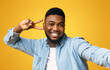 © Prostock-studio - Say Cheese. Joyful black millennial guy showing peace sign while taking selfie, yellow studio background with free space