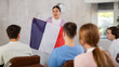 © JackF - Positive friendly young female tutor giving lecture to group of students during educational class, holding national flag of France and talking about history of country