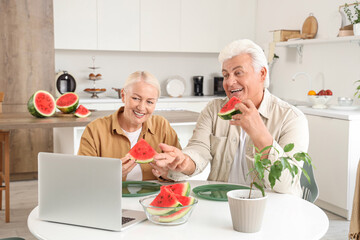 Canvas Print - Mature couple with slices of fresh watermelon video chatting at table in kitchen