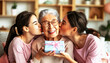 © sennauli - Two Young Women Kissing a Grandmother Holding a Present