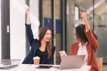 Two diverse businesswomen excitedly raise their hands happy about good news about collaboration on digital tablets and laptops in the office.