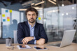 © Liubomir - Confident businessman in a modern office environment, seated at a desk with laptop and tablet, exuding professionalism and determination.