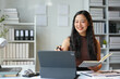 © Phimwilai - Young woman working on laptop in office Asian businesswoman sitting and taking notes in a notebook Thinking of ideas at your desk Beautiful independent woman working online with paperwork.