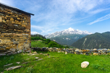 Wall Mural - The medieval stone basilica stands on a green field surrounded by a stone fence. Bright blue sky and snow-capped Mount Tetnuldi in the background