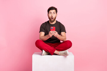 Poster - Full size photo of impressed man dressed brown t-shirt sit on cube hold smartphone astonished staring isolated on pink color background