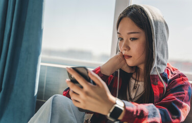 Beautiful serious Asian teenage girl sitting by the window and looking at the smartphone screen, portrait