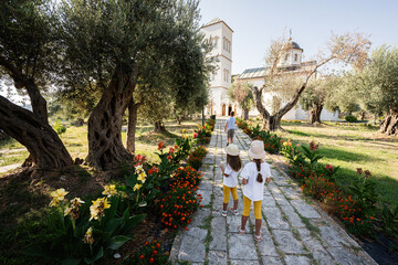 Children exploring a scenic garden path leading to a historic church on a sunny day, surrounded by olive trees and colorful flowers in Ulcinj, Montenegro.