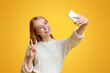© Prostock-studio - Smiling teenage girl taking selfie on smartphone, showing peace gesture, orange studio background with copy space
