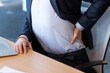 © Liubomir - Business professional sitting in office chair showing signs of back pain. Person dressed in suit experiencing discomfort, highlighting health issues related to sitting posture during work hours.