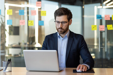 Poster - Businessman concentrating on laptop work in modern office setting, surrounded by colorful sticky notes on glass wall. Portrays focus, productivity, and analysis in business environment.