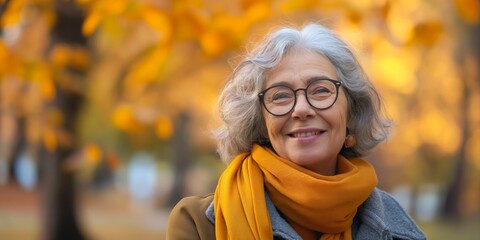 Wall Mural - A woman wearing glasses and a yellow scarf is smiling. She is standing in a park with trees in the background