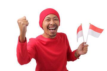 Excited Indonesian man in a red long sleeve T-shirt and beanie holds the Indonesian flag and raises his fist in celebration of Indonesian Independence Day on August 17, isolated on a white background