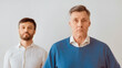 © Prostock-studio - Serious Senior Man And His Middle Aged Son Standing Looking At Camera Against White Wall. Panorama, Selective Focus