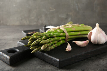 Wall Mural - Board with bunch of fresh green asparagus stems and garlic on grey textured table, closeup