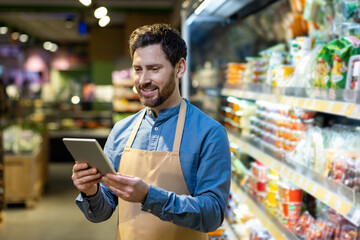 grocery store employee wearing apron stands in supermarket aisle using tablet. man checks inventory 