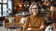 © aicandy - Confident middle-aged woman with glasses, wearing a brown sweater, standing with arms crossed in a warmly lit home living room with cozy decor and modern furnishings
