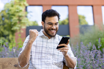 Wall Mural - Close-up photo of a happy young Indian man enjoying victory and success, sitting outside on a bench and reading messages and news on his phone, showing a winning gesture