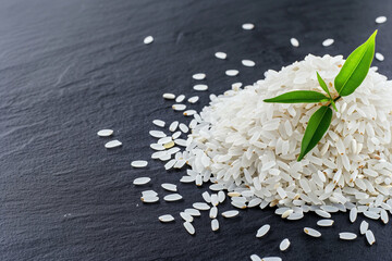 Wall Mural - Rice pile with green leaves on a dark stone background. Fresh food texture showing uncooked rice grains scattered around. Organic rice with natural elements