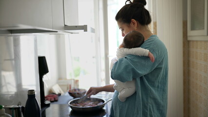 Mom in blue shirt holding baby while stirring food in a pan on the stove, illuminated by natural light through the window, showcasing multitasking, care, and balancing parenting with daily chores