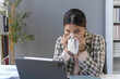 © amnaj - Businesswoman with cold or flu sneezing in tissue at office desk while working on laptop