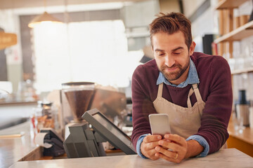 Fototapete - Businessman, barista and cafe with phone for online order, service or social media at coffee shop. Happy man, waiter or business owner with smile on mobile smartphone for review at indoor restaurant