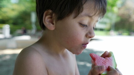 Close-up of young boy holding and eating a slice of watermelon, shirtless and focused, enjoying a refreshing summer snack, outdoor fun in the park, healthy and hydrating, playful day