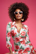 © EVISUAL - Stylish and Sunny: Beautiful woman posing in nice flower pattern dress and sunglasses on a pink background. Fashion photo with afro hairstyle. generative AI
