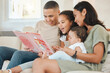 © peopleimages.com - Parents, children and story book on sofa in home with smile, reading and listening for bonding in living room. Father, mother and daughter with baby, happy and connection at family house in Colombia