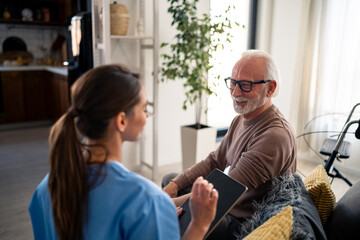 A joyful female nurse in blue scrubs engages with an elderly male patient in his home, discussing health matters while seated comfortably.